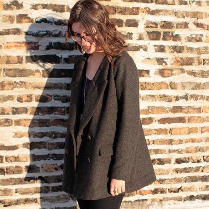 Person wearing a gray wool coat with oversized lapels and shawl collar over a dark top, standing against a brick wall