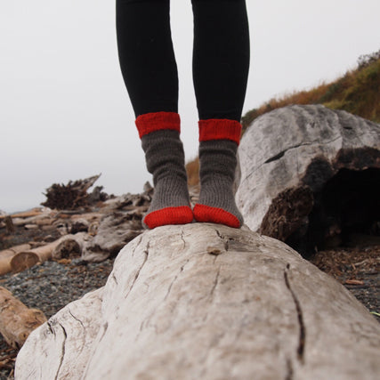 Person in black leggings and hand-knit gray socks with red striped cuffs standing on a driftwood log at a rocky beach