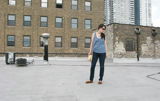 Woman in sleeveless chambray tank and dark jeans standing on an urban rooftop with brick buildings in background