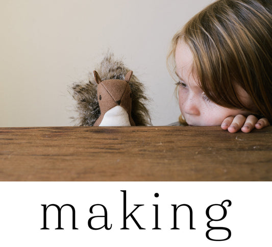 Child peeking over wooden table edge at a handmade felt and fur stuffed squirrel toy, with "making" text below