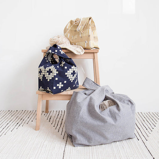 Three Stowe Bags in cream woven, navy geometric, and gray linen on a wooden step stool with yarn and patterned rug