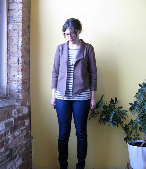 Person wearing a taupe knit cardigan over a striped tee with dark jeans, standing against a yellow wall next to exposed brick