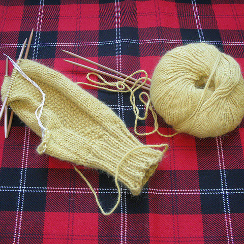 Cream-colored stockinette knit mittens in progress with yarn and needles on red and navy plaid fabric