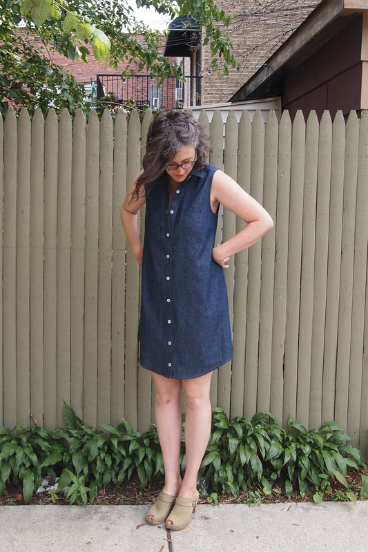 Woman wearing a sleeveless dark denim Alder Shirtdress with collar, chest pocket, and white buttons, wearing tan clogs