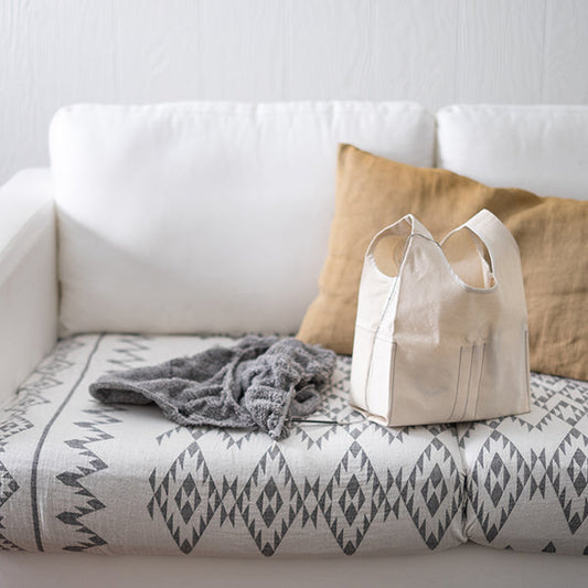 Stowe Bag in natural canvas on a white couch with a geometric print blanket, gray knitting project, and tan linen pillows