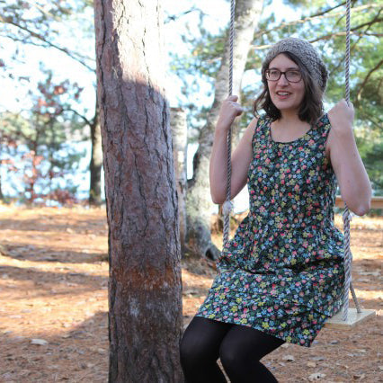 Woman wearing floral print sleeveless Edenham Liberty print dress with black tights on a swing in a forest setting
