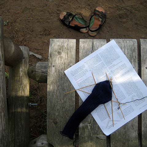 Navy knit sock on double-pointed needles resting on printed pattern on a weathered wood surface with sandals nearby
