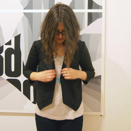 Person in a black Morris Blazer over cream textured tank with turquoise necklace in front of a black and white graphic artwork