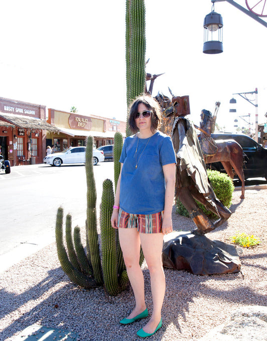 Woman in blue tee and ikat Maritime Shorts with turquoise flats standing next to tall cacti at a desert town storefront