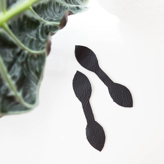 Two black leather zipper pull pieces cut to shape for Portside Dopp Kit, beside green plant leaf on white background