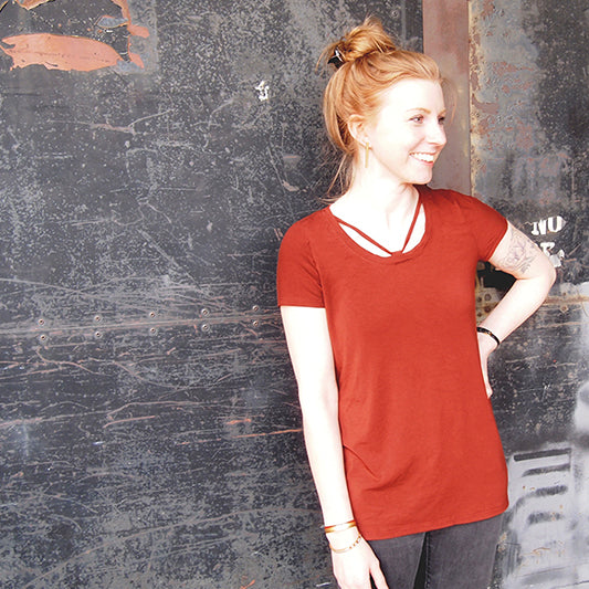 Person wearing a rust red Lark Tee with cross-strap neckline variation and short sleeves, against a weathered metal wall