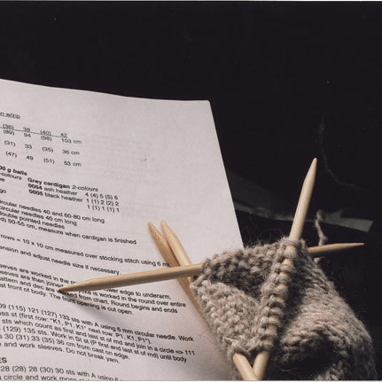 Gray wool knitting in progress on wooden needles resting on printed pattern instructions with ribbed sleeve sections visible