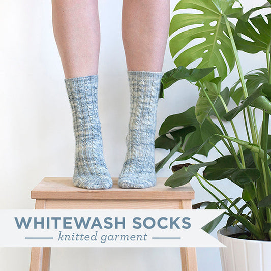 Handknit Whitewash Socks in blue and cream cable knit, displayed on wooden stool next to monstera plant, with text overlay