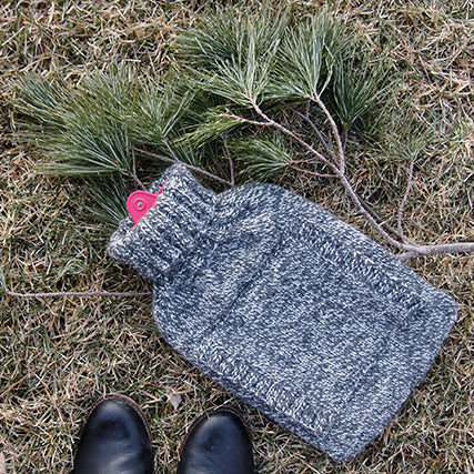 Knit hot water bottle cozy in blue heather with pink toggle closure on dried grass with evergreen branches and black boots