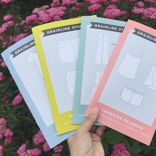 Hand holding four Grainline Studio pattern envelopes in blue, yellow, mint, and pink fanned out against pink flowering shrubs