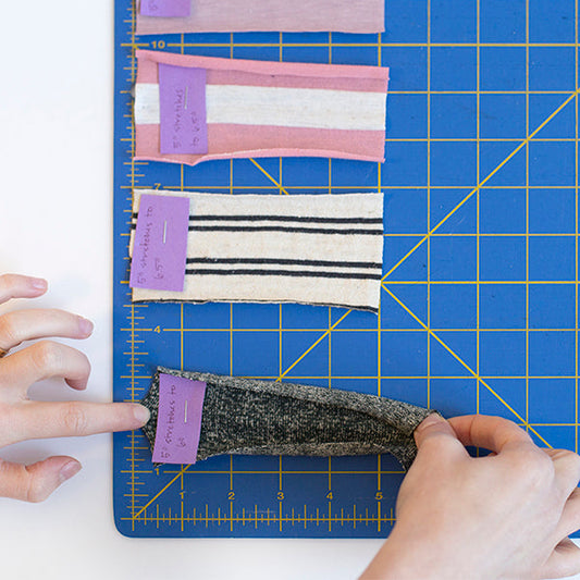 Hands measuring stretch percentages on knit fabric swatches on a blue cutting mat, with purple labels noting results