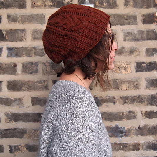 Side profile of person wearing the Bray cable knit slouchy hat in rust, paired with a gray sweater, against a brick wall