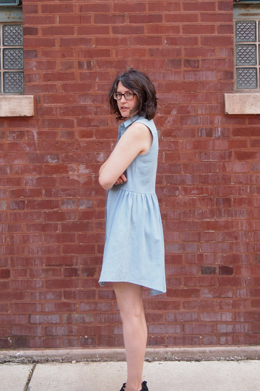 Side view of woman wearing a sleeveless light-wash denim Alder Shirtdress with collar, gathered skirt, and shirt-tail hem
