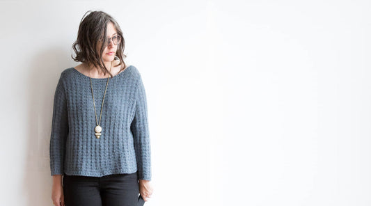 Gray ribbed knit sweater with long sleeves and wide crewneck, worn with black pants and a long pendant necklace