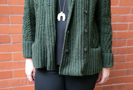 Handknit forest green Exeter Cardigan with shawl collar and double-breasted buttons, worn over black top against brick wall