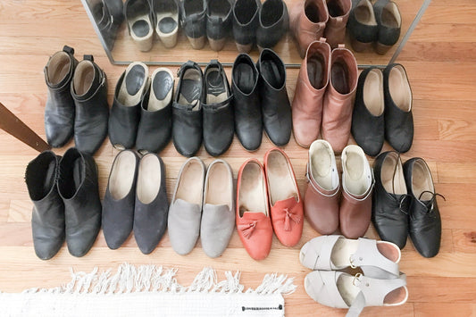 Overhead view of 13 pairs of shoes including boots, loafers, and clogs in black, gray, rust, and tan on wood floor