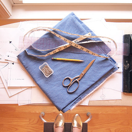 Flat lay of sewing supplies on pattern pieces: blue fabric, measuring tape, scissors, pins, and weights on ironing board