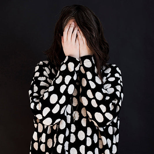 Person wearing a black and white jacquard polka dot long-sleeve sweatshirt, hands covering face against dark background