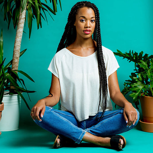 Person in white Penny Raglan tee and jeans, seated cross-legged against a turquoise background surrounded by potted plants