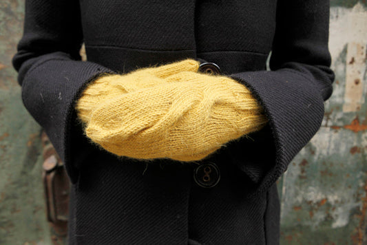 Hands in golden cabled handknit mittens tucked under navy wool coat sleeves with industrial background