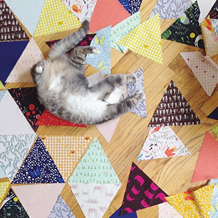 Gray tabby cat lying on colorful triangle quilt pieces arranged on a wood floor, showing a variety of prints and colors
