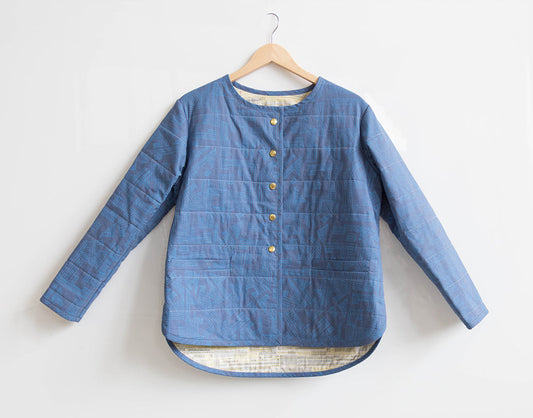 Tamarack Jacket in blue geometric print with crew neckline, gold snap closures, and cream lining at hem, on a wooden hanger