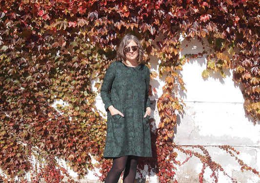 Farrow Dress in green and black print with long sleeves and black tights, standing in front of an autumn ivy-covered wall
