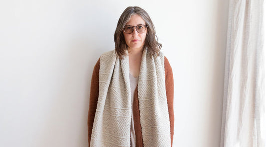 Person with gray hair and glasses wearing rust cardigan and oversized cream knit scarf, standing against a white wall