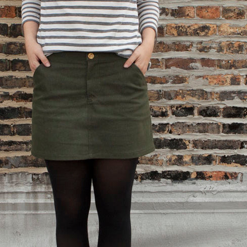 Person wearing striped long-sleeve shirt and olive green denim Moss Skirt with black tights against brick wall