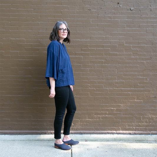 Person wearing blue silk noil Luna Jacket by Papercut Patterns with black jeans and mules, against a brick wall