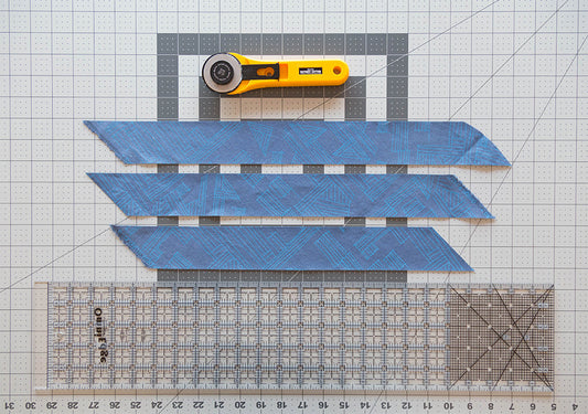 Three blue geometric bias strips cut for the Tamarack Jacket on a gridded cutting mat with rotary cutter and quilting ruler