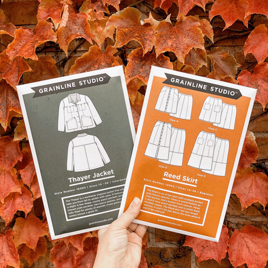 Hand holding Grainline Studio Thayer Jacket and Reed Skirt pattern envelopes, sizes 14-30, against autumn leaves background