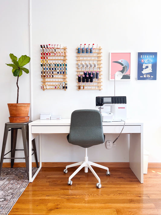 Organized sewing studio with IKEA Micke desk, BERNINA serger, gray chair, two wooden thread racks, and fiddle leaf fig plant