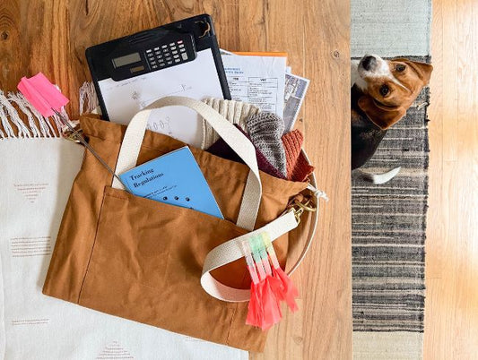 Tan canvas Town Bag filled with AKC tracking handbook and supplies on wood floor, with beagle peeking in from edge