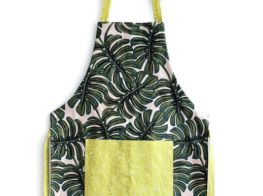 Grainline Apron with green and cream tropical leaf print body, yellow polka dot pocket and straps, on white background