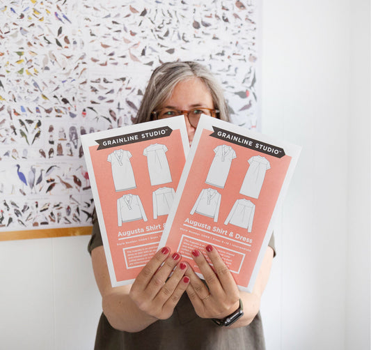 Person holding two Grainline Studio Augusta Shirt & Dress pattern envelopes with coral backgrounds and garment sketches