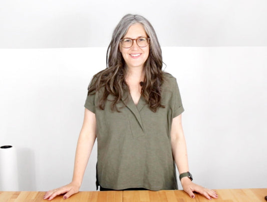 Person wearing olive green short-sleeved Augusta Shirt with V-neckline, standing at wooden table in front of white wall