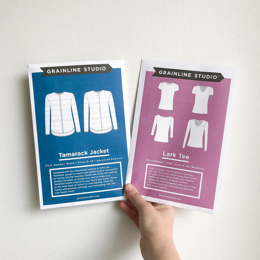 Hand holding Grainline Studio Tamarack Jacket and Lark Tee pattern envelopes in blue and mauve, showing garment sketches