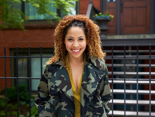 Woman wearing mustard Augusta Top under green camo print Cortland Trench, standing by brick building and iron fence