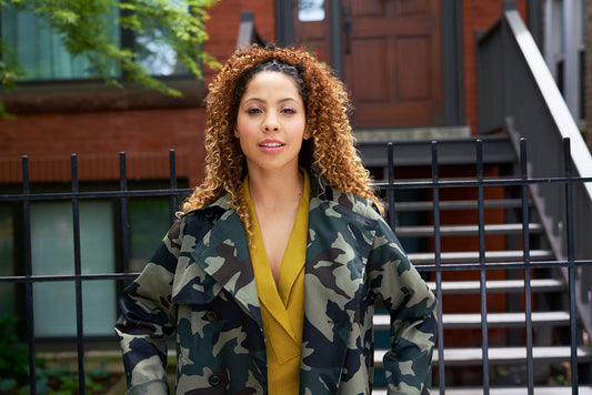 Woman wearing mustard Augusta Top under green camo print Cortland Trench, standing by brick building and iron fence
