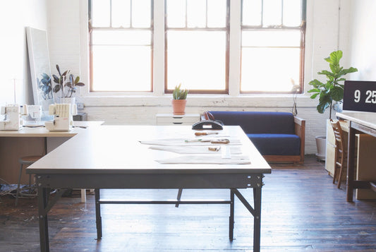 Grainline Studio workspace with large cutting table, pattern pieces, sewing machines, plants, and large industrial windows