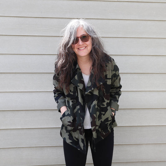 Woman wearing unbuttoned hip-length camo Cortland Trench with storm flap, over white Penny Raglan and black jeans