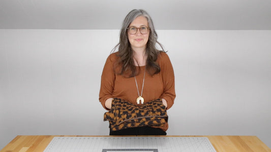 Person wearing rust Hemlock Tee holding rust and black geometric print fabric at wood table with cutting mat during sew-along