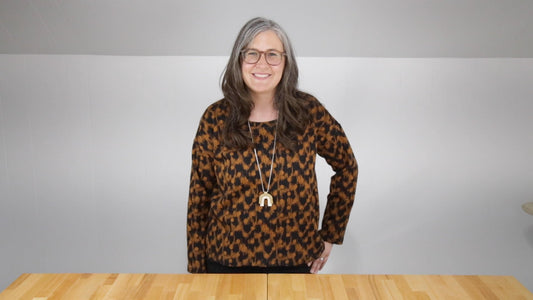 Person wearing rust and black geometric print Hemlock Tee with long sleeves and gold pendant necklace, standing at wood table