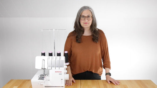 Person wearing rust Hemlock Tee standing at wood table next to a white Bernina serger with black and pink thread spools